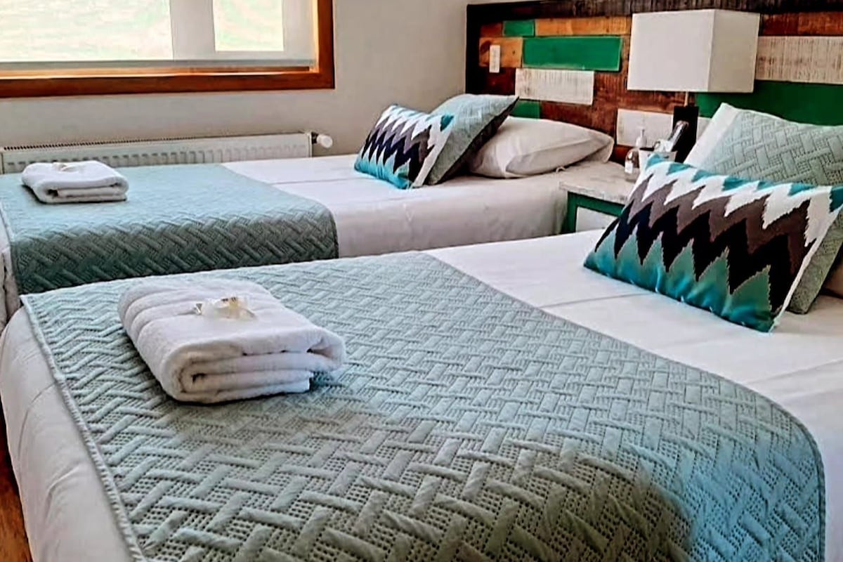 Une chambre d’hôtel avec deux lits, chacun recouvert d’une courtepointe matelassée verte et agrémenté d’oreillers, dotée d’une tête de lit en bois et d’une fenêtre donnant sur l’extérieur.