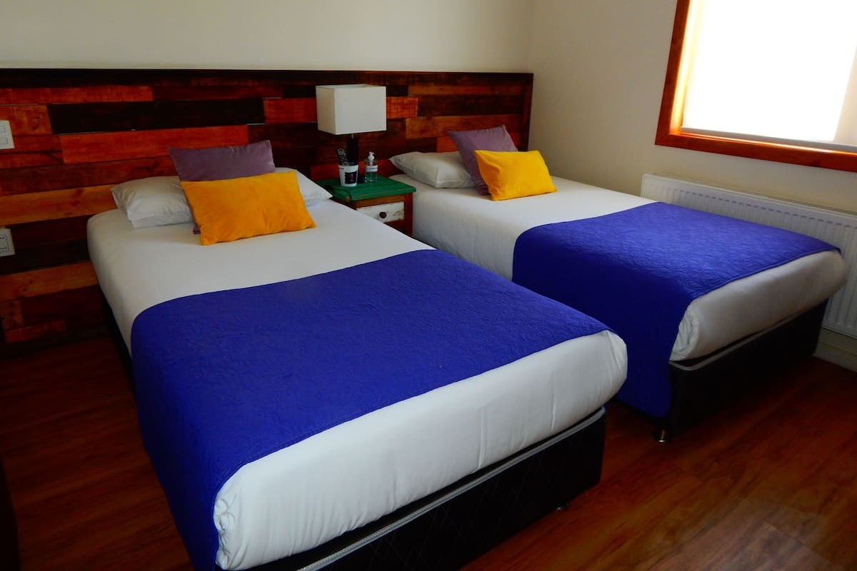 Ein Hotelzimmer mit zwei Betten, jeweils mit blauer Bettdecke und weißen Laken. Es gibt ein hölzernes Kopfteil, einen Nachttisch mit Lampe und ein Fenster, das natürliches Licht hereinlässt.