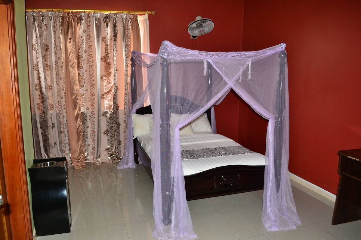 Ein Schlafzimmer mit einem Himmelbett, einem Ventilator am Nachttisch und einem Vorhang mit Blumenmuster.