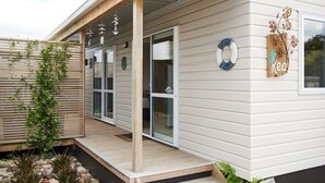 A modern house with a covered porch, sliding glass doors, and a small garden area with plants and a wooden fence. - Kea Cabin - beach/restaurants/supermarket all within 5 min walk. (Orewa Beach)