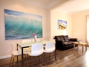 A modern living room with a dining area, featuring a large wave painting, a brown leather sofa, and a small table with a vase of flowers. - CLASSBEDROOM EIXAMPLE FREE 211, 1 1 (Barcelona)