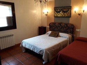 A bedroom with a bed, a nightstand, a radiator, a window with curtains, and a wall-mounted light fixture.