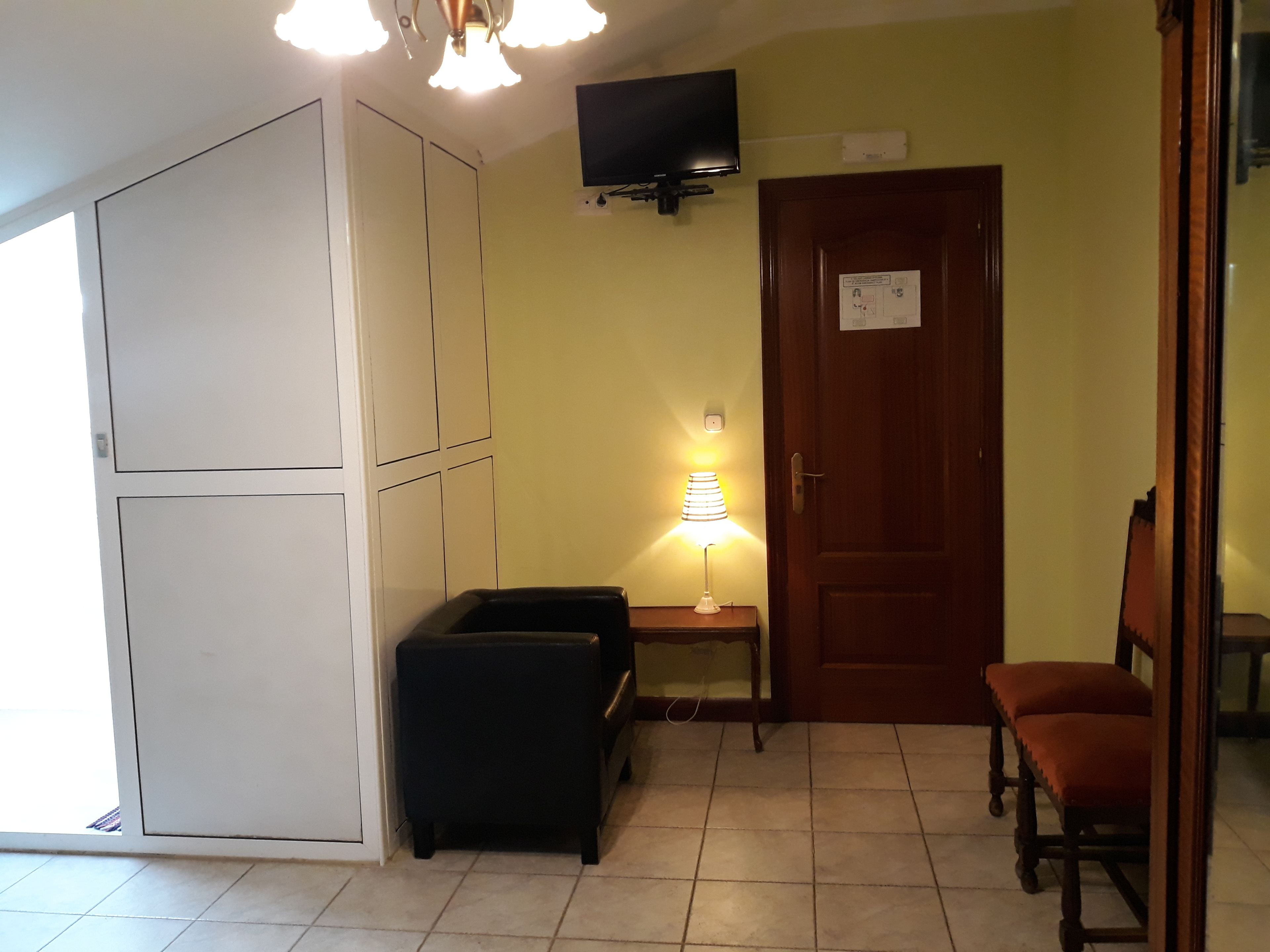 A compact room with a bed, a small table with a lamp, a TV mounted on the wall, and a door with a sign.