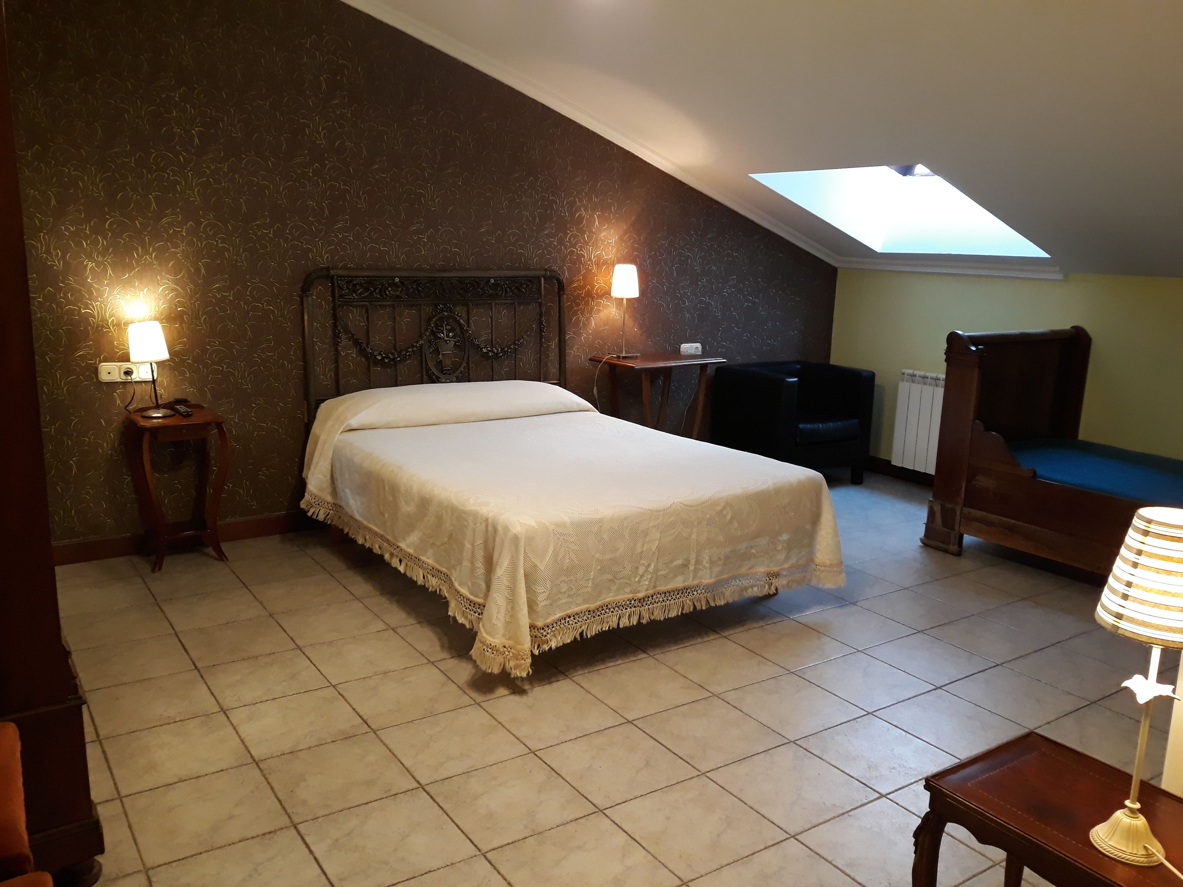 A bedroom with a bed, two bedside lamps, a chair, a small table, and a skylight.