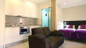 A modern hotel room with a kitchenette, a sofa, two beds with purple bedding, and a bathroom with a blue shower area. - CLASSBEDROOM BARCELONA BEACH 2 APARTMENT (Barcelona)