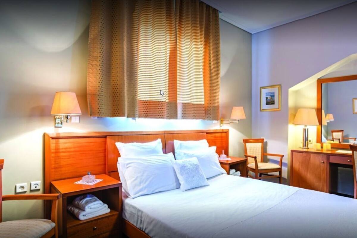 A hotel room with a bed, bedside tables, a desk, a chair, a wardrobe, and a large window with curtains.