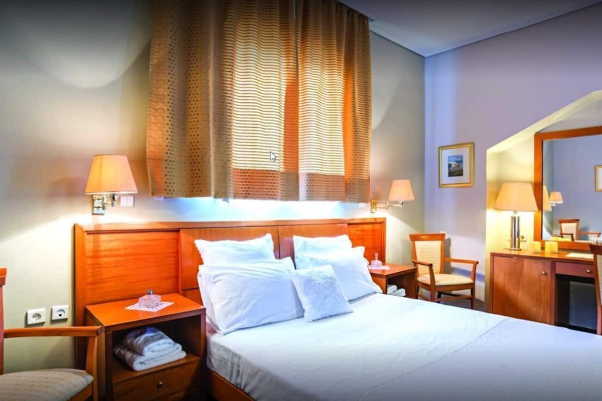 A hotel room with a bed, bedside tables, a desk, a chair, a wardrobe, and a large window with curtains.