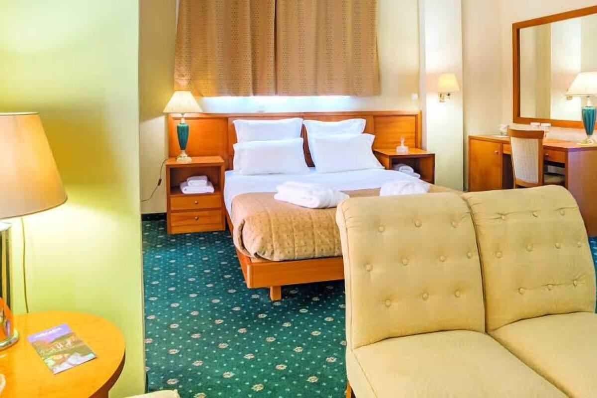 A hotel room with a bed, two bedside tables with lamps, a chair, a desk with a mirror, and a window with curtains.