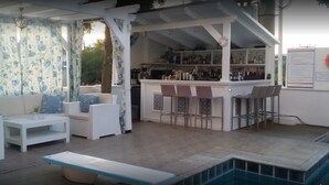 A covered outdoor seating area with white furniture, a pool, and a bar with shelves of bottles. - Hotel Philippos (Livadia)