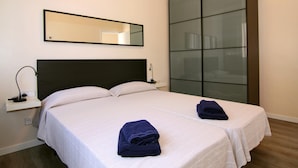 Un letto matrimoniale con biancheria da letto bianca e due asciugamani blu scuro piegati. Si intravede un comodino con una lampada e una presa di corrente. Sopra il letto è appeso uno specchio grande, e a destra si trova una porta scorrevole in vetro.