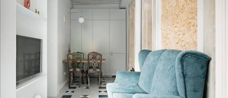 A modern living room with a blue sofa, a dining table with chairs, and a TV on the wall.