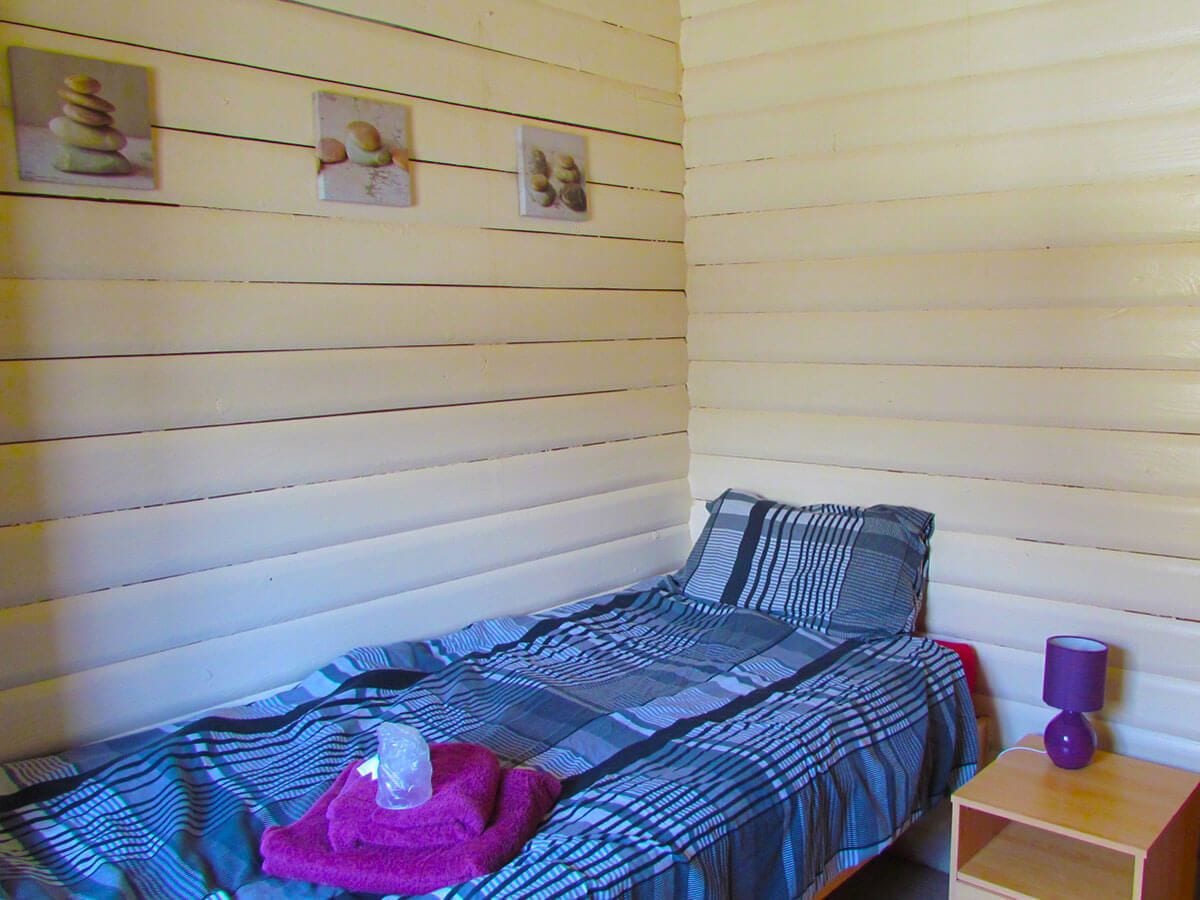 Une petite chambre avec un lit simple, une couvre-lit à rayures, un oreiller violet et une petite table de chevet avec une lampe.