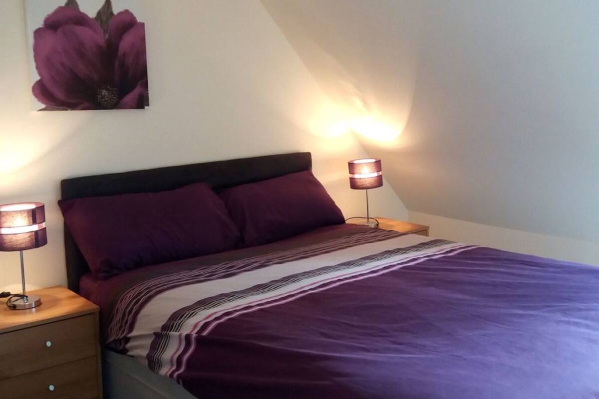 A bedroom with a bed, two bedside lamps, a nightstand, a large purple flower painting on the wall, and a slanted ceiling.