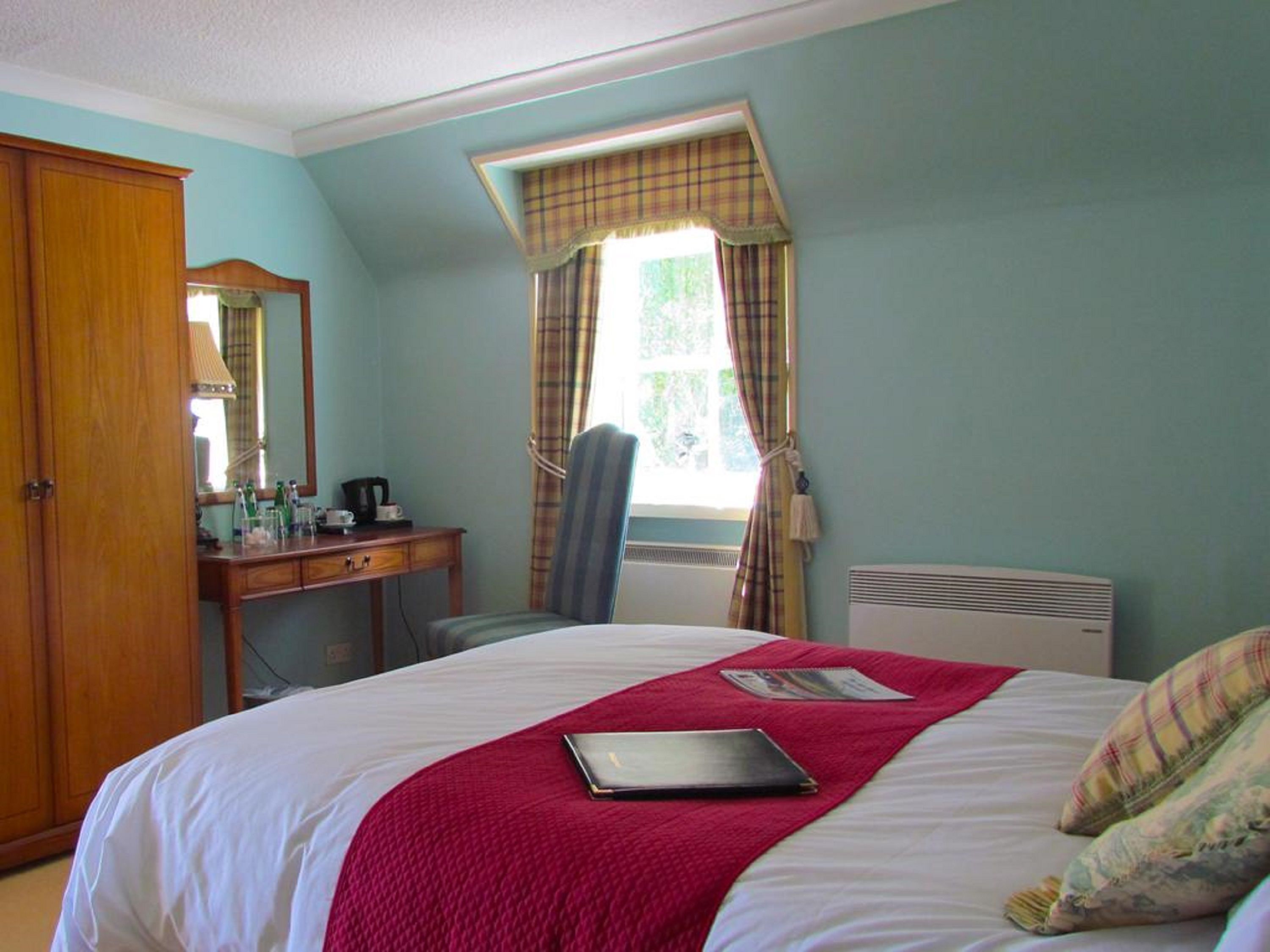 A bedroom with a bed, a desk, a chair, a window with curtains, and a radiator.