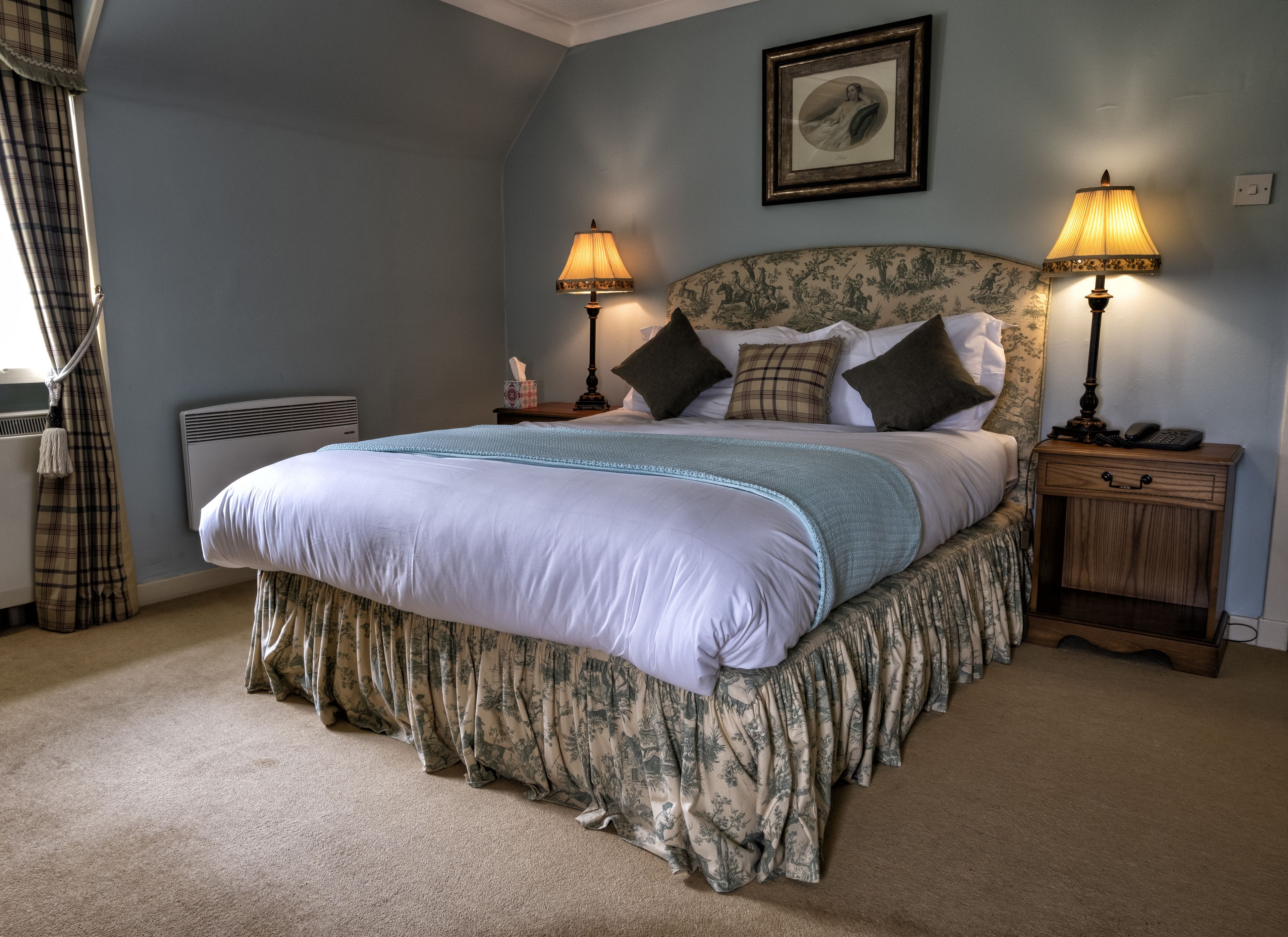 A neatly made bed with a floral bed skirt, two bedside lamps, a framed picture on the wall, and a nightstand with a phone and lamp.