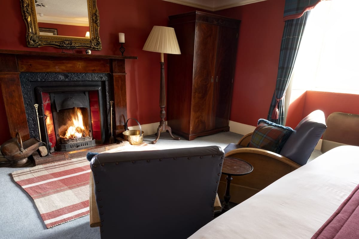 A hotel room with a fireplace, a bed, a chair, a lamp, and a window with curtains.