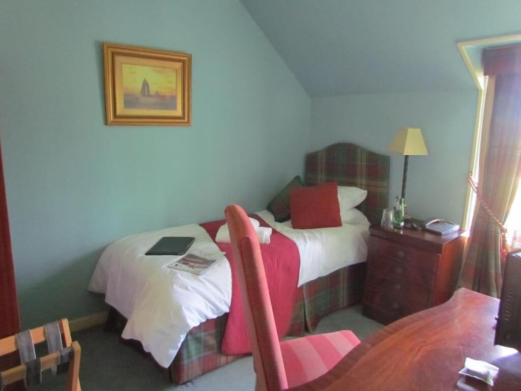Un dormitorio con cama, mesita de noche, silla, lámpara y un cuadro en la pared.