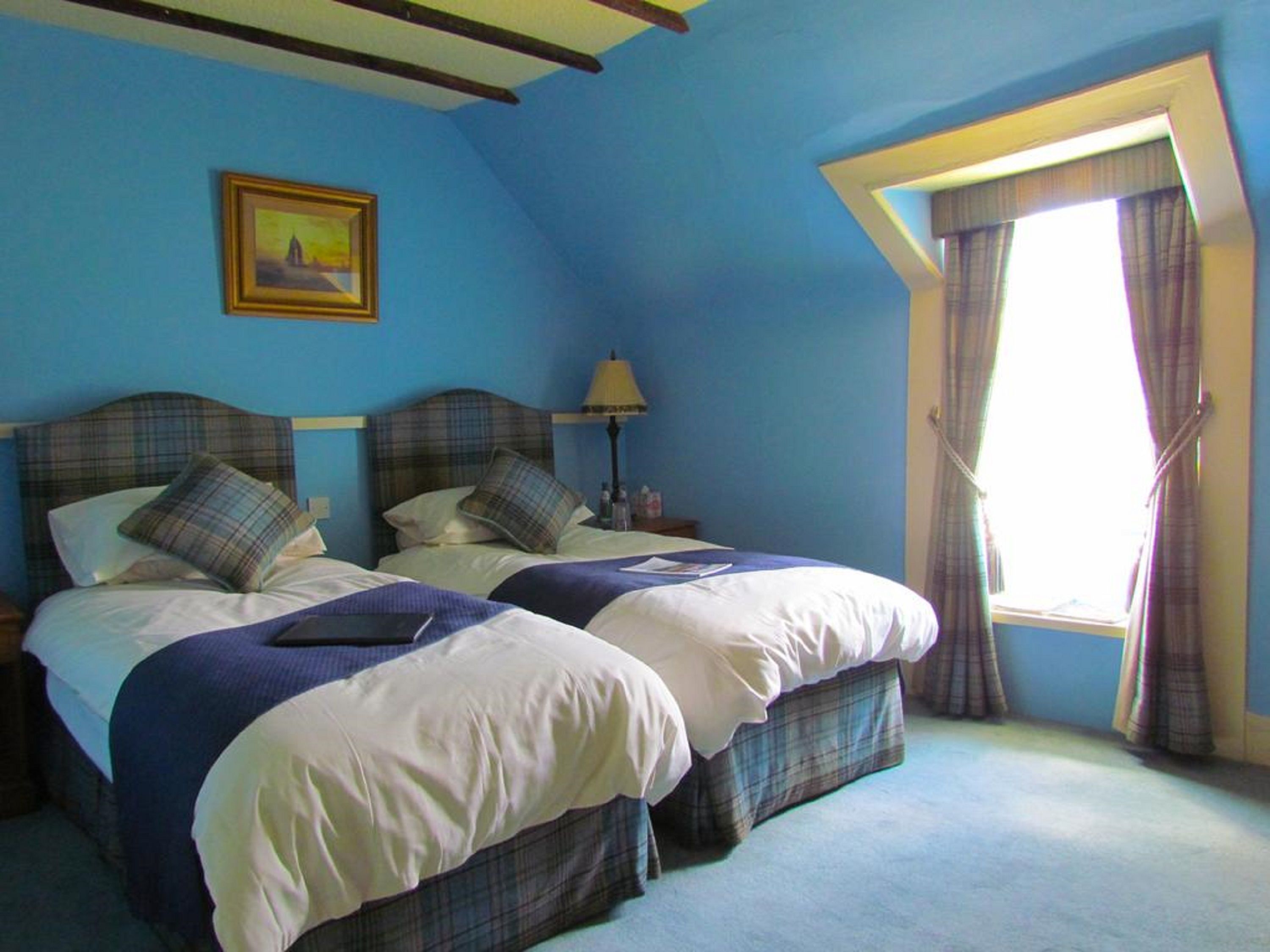 Une chambre avec deux lits, un tableau au mur, une lampe et une fenêtre avec des rideaux.