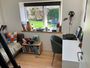 Une pièce comprenant un lit, un bureau avec un ordinateur, une chaise, une bibliothèque et une fenêtre donnant sur un jardin.