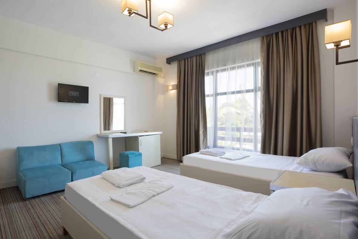 Une chambre d’hôtel avec deux lits, un bureau, un canapé bleu et une grande fenêtre avec des rideaux.