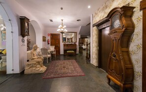 A hotel lobby with a large wooden grandfather clock, a statue, a chandelier, and a patterned carpet. - Alda Vía León (León)
