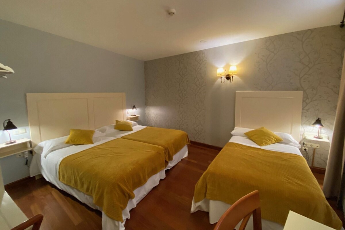 A hotel room with two beds, each with a white headboard and a yellow blanket. There are bedside lamps, a small table with a lamp, and a chair. The room has a patterned wallpaper and a wooden floor.