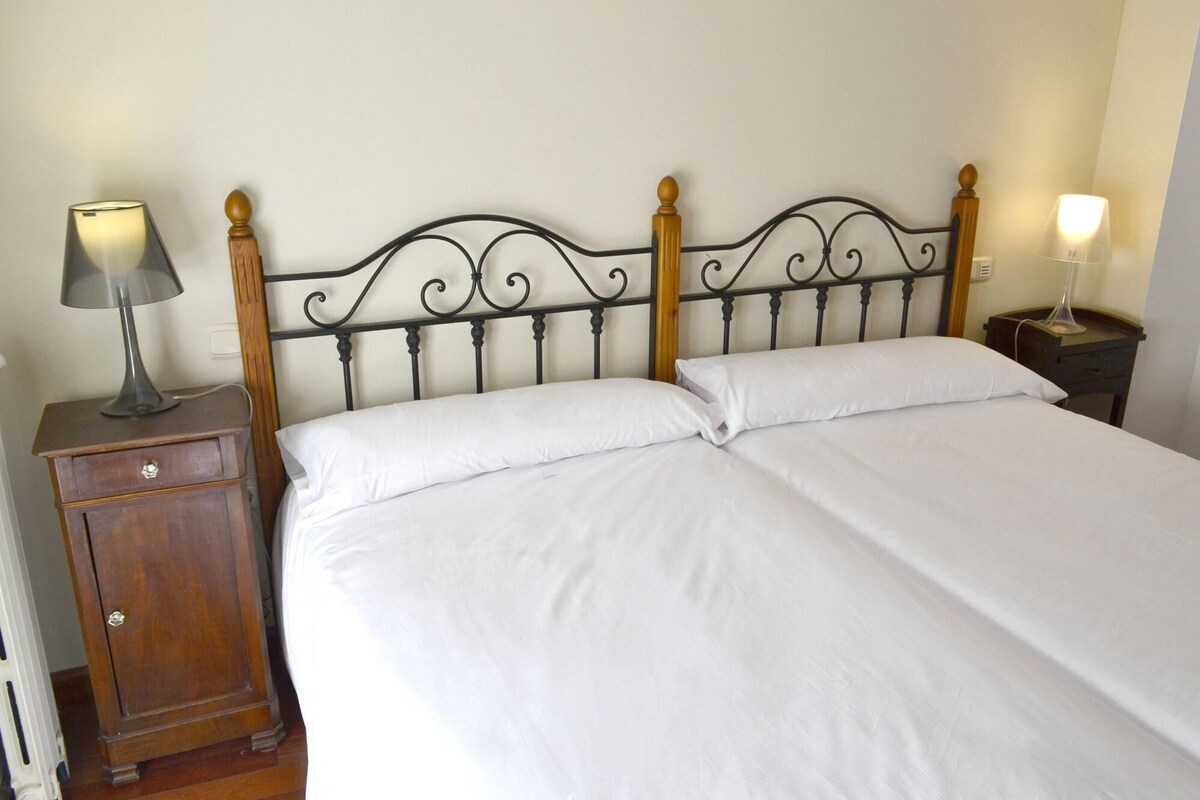 A neatly made bed with white linens, a wooden nightstand with a lamp, and a wall-mounted light fixture.