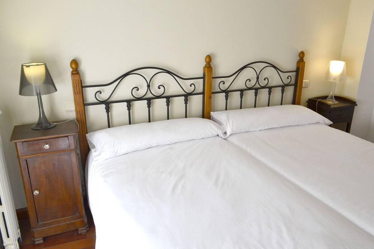 A neatly made bed with white linens, a wooden nightstand with a lamp, and a wall-mounted light fixture.