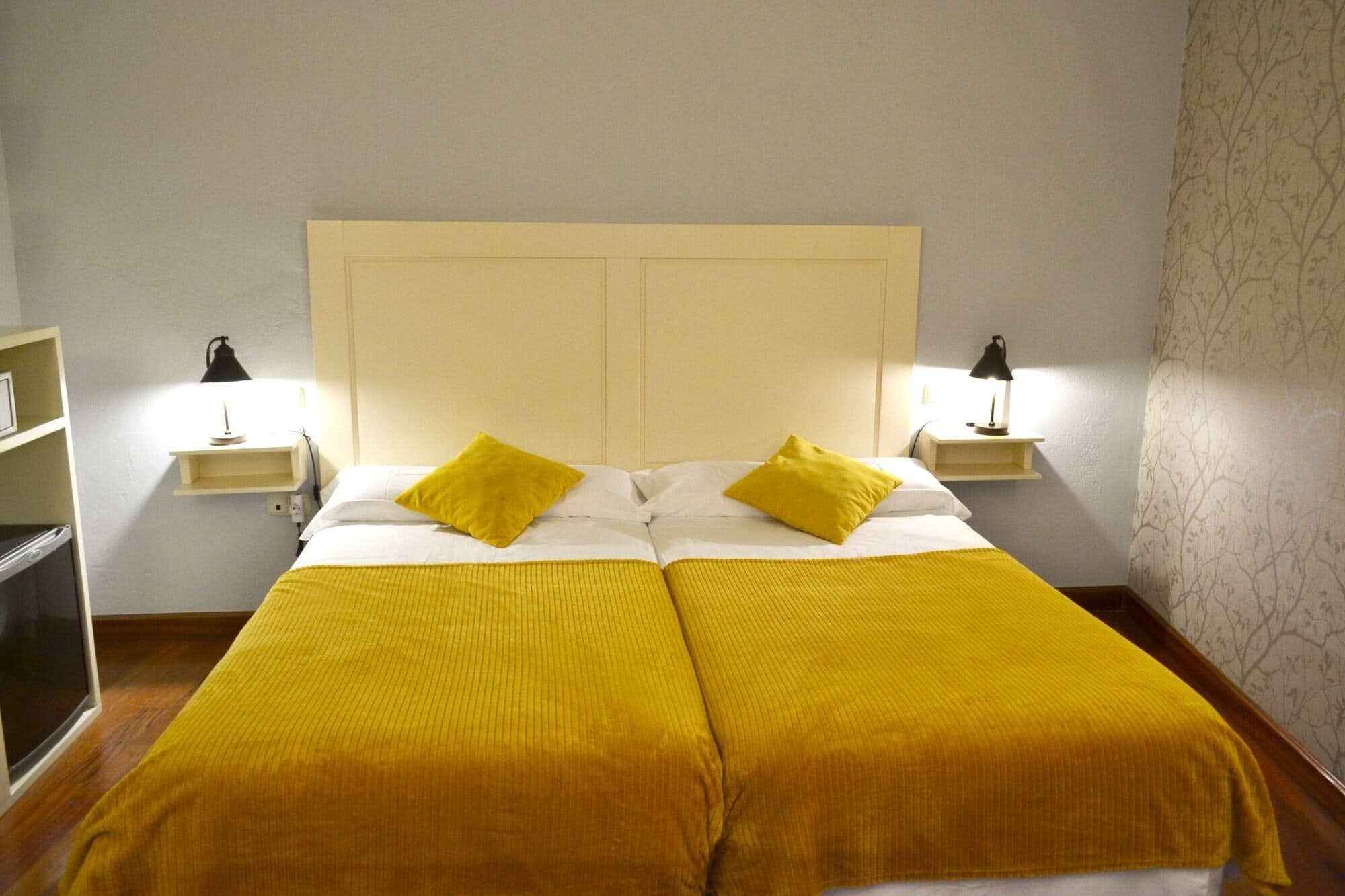 A double bed with two yellow pillows, a white headboard, and two bedside lamps.