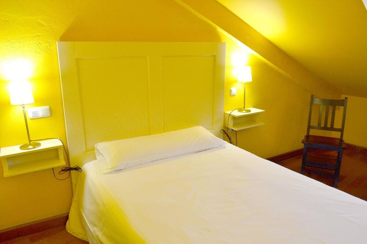 A single bed in a room with yellow walls and a sloped ceiling. There are two bedside lamps on white nightstands. A wooden chair is visible in the corner.