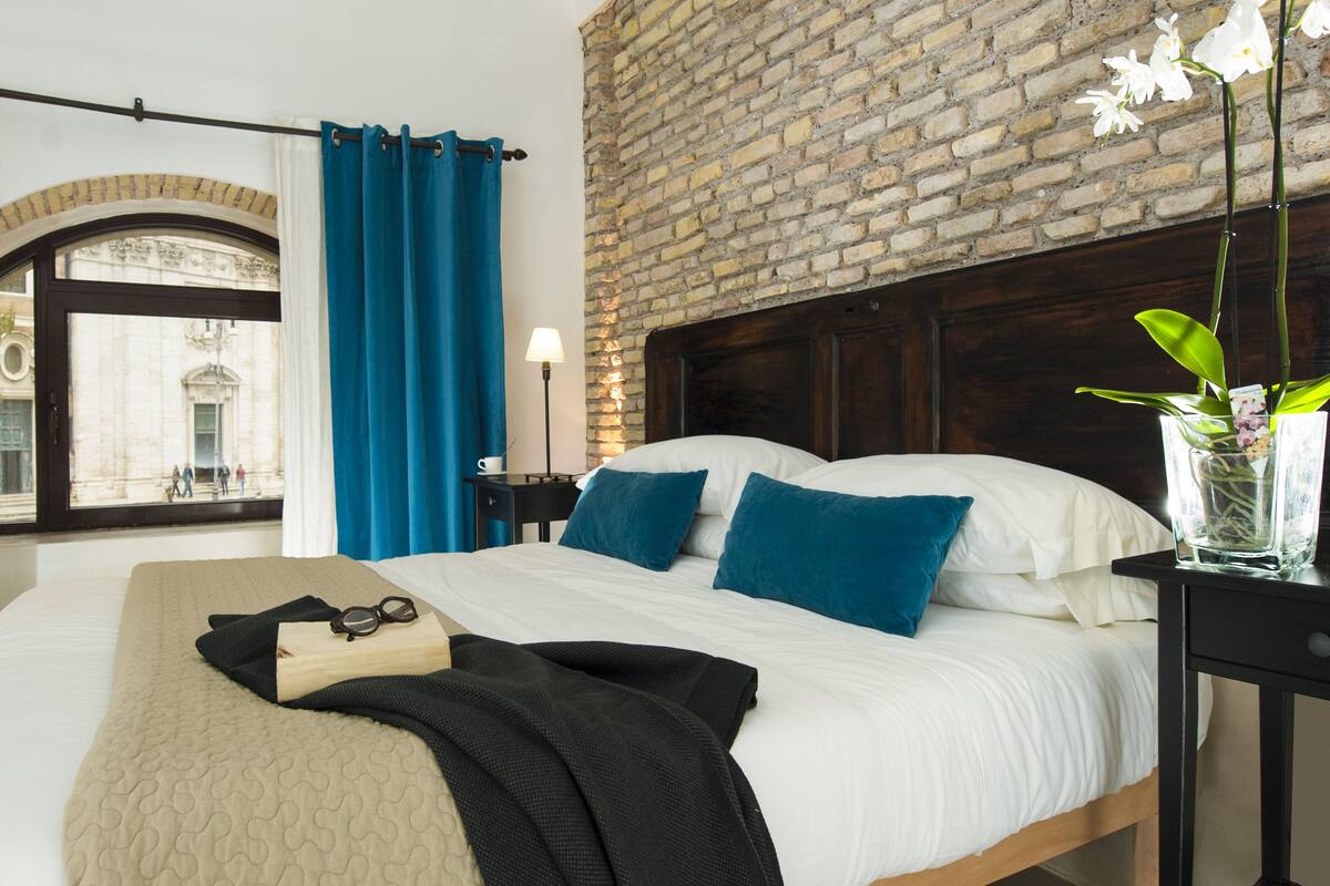 A hotel room with a brick wall, a bed with white and blue bedding, a nightstand with a lamp, and a window with blue curtains.