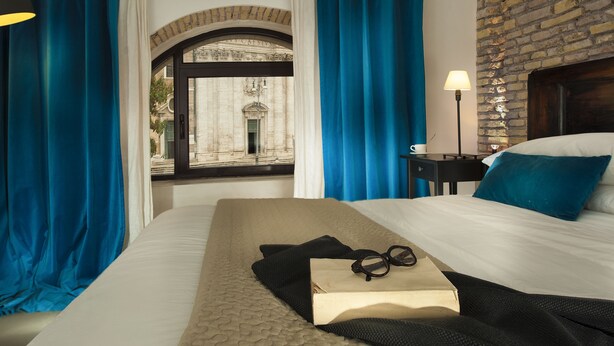 A hotel room with a bed, a window with blue curtains, a lamp, and a view of a building outside. - Corso Vittorio Apartments (Rome)