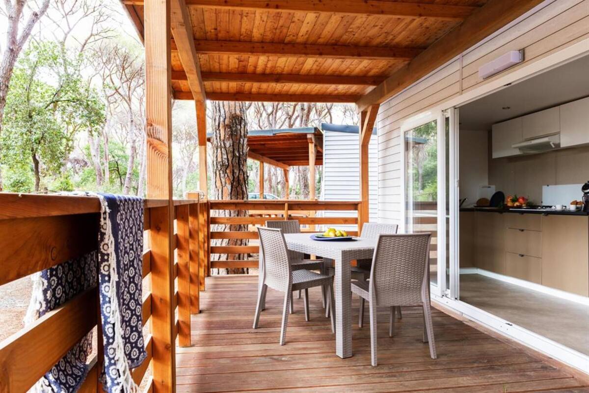 Eine Holzterrasse mit Tisch und Stühlen, einem überdachten Bereich und Blick auf eine Küche durch Glastüren.