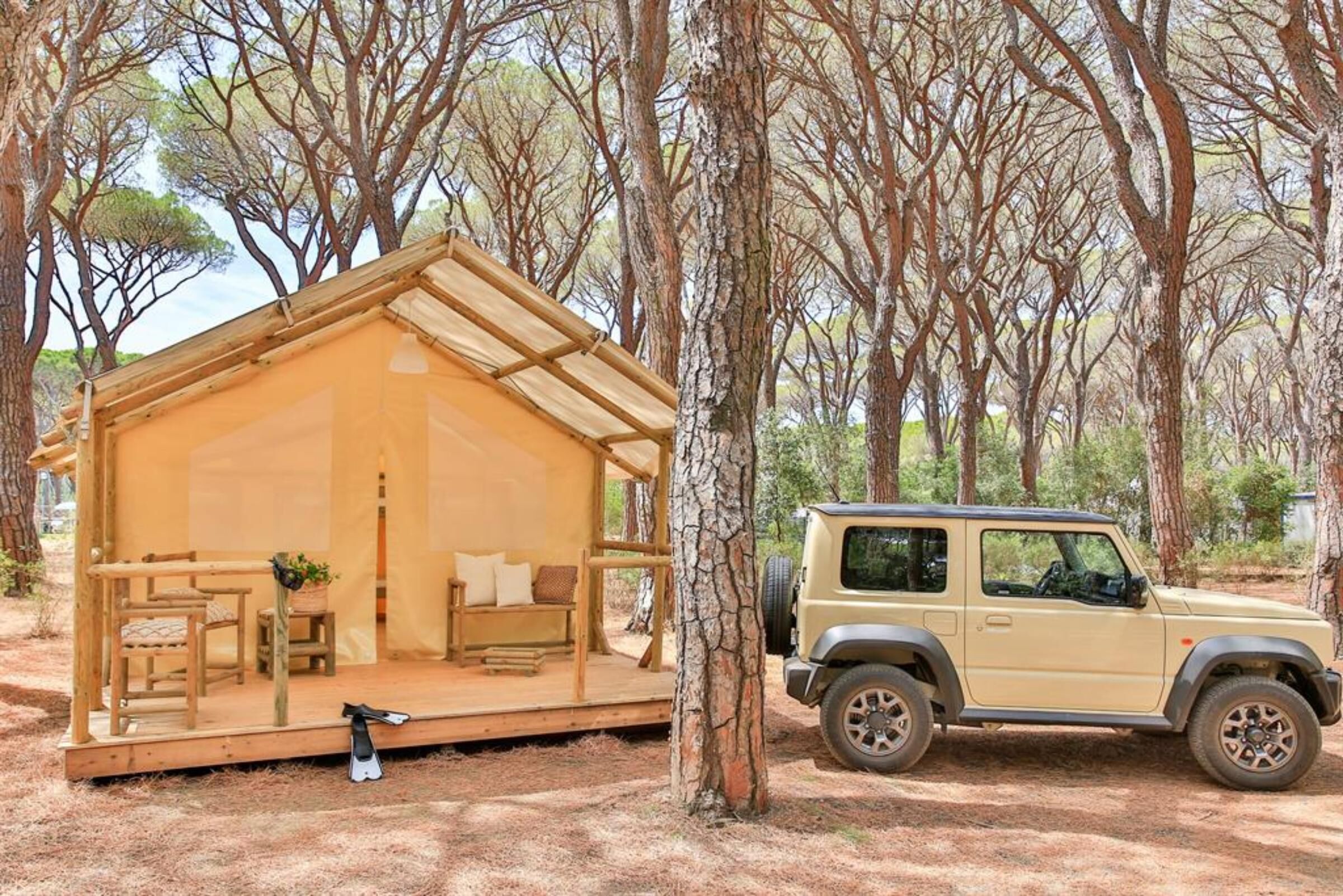Una pequeña estructura de madera con un porche y una zona de estar, un vehículo estacionado cerca y un fondo boscoso.