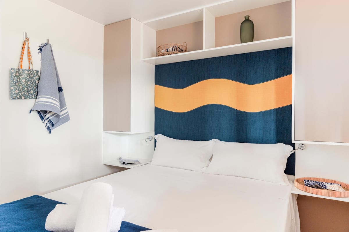 Ein modernes Hotelzimmer mit einem Bett, einem Kopfteil mit wellenförmigem Design, einer kleinen Ablage mit einer Vase und einem Korb.