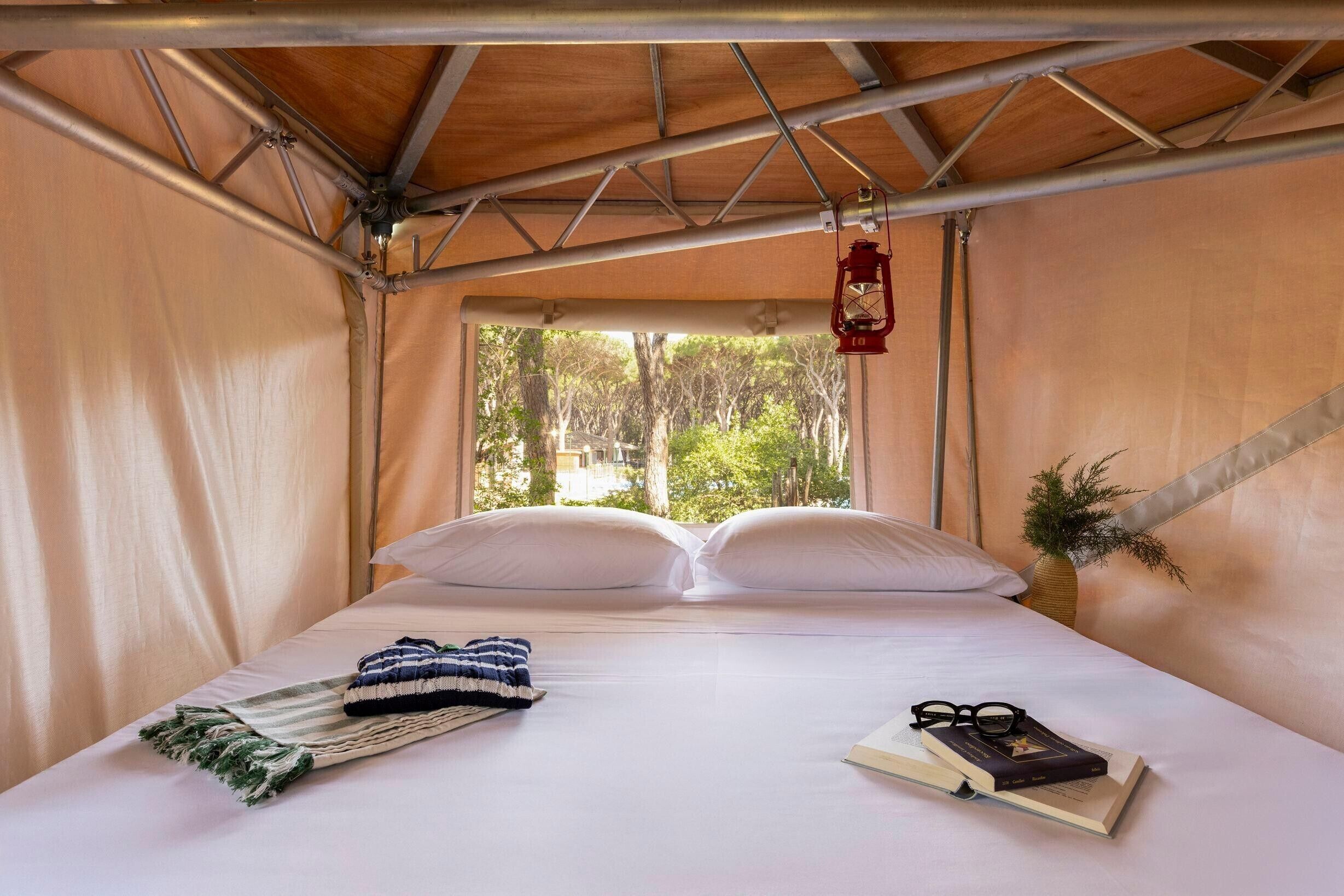 A bed with white linens, a striped blanket, a book, and glasses, a red lantern, and a view of trees through a window.