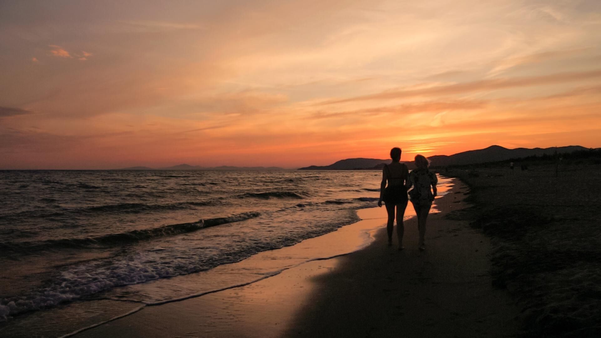 Eine Szene am Strand, zwei Personen spazieren während des Sonnenuntergangs entlang.