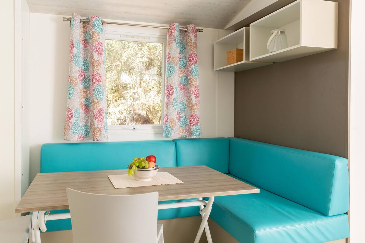 Ein kleiner Raum mit einem senffarbenen Sofa, einem Tisch mit Obstschale und einem Fenster mit gemusterten Vorhängen.