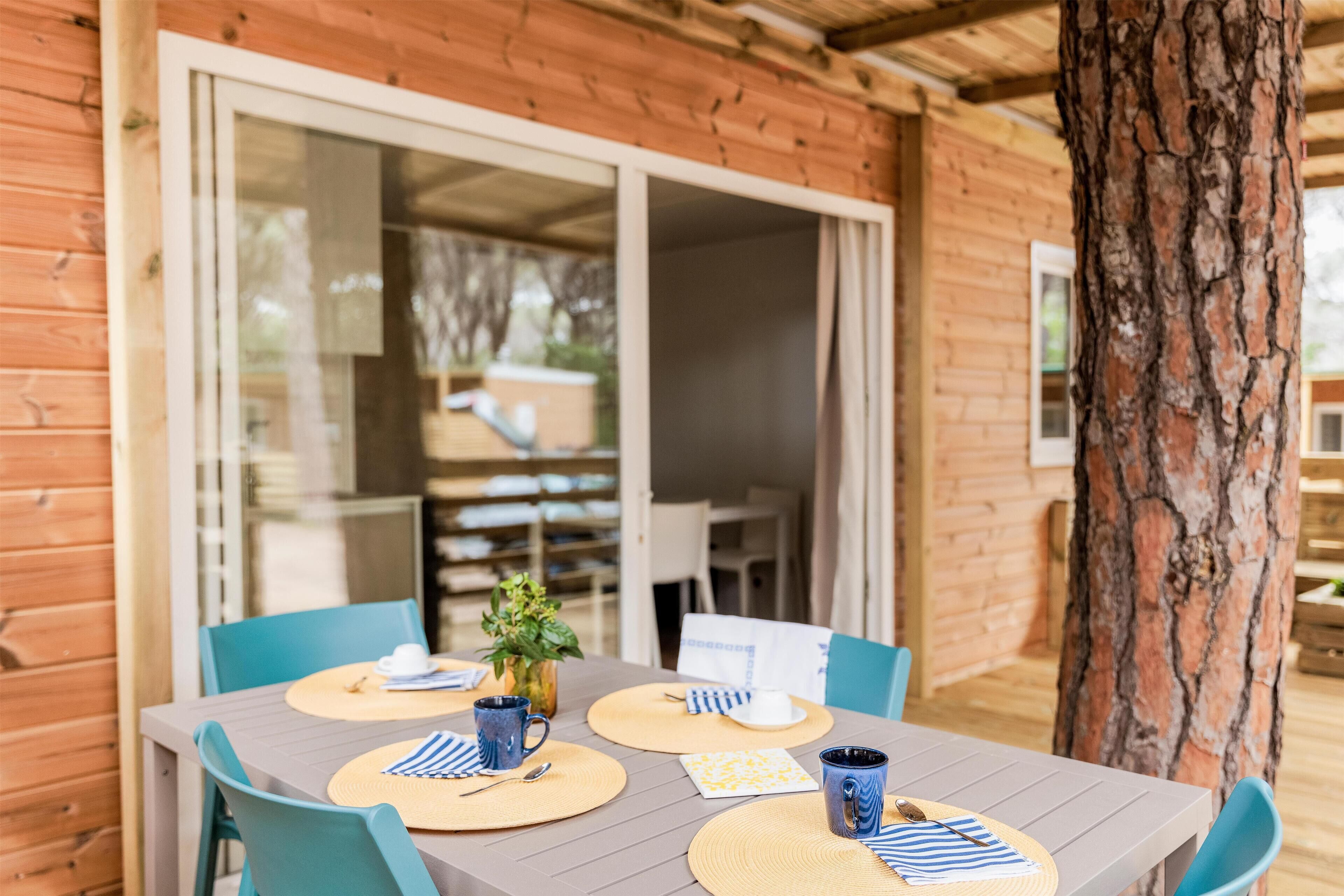 Une cabane en bois avec une table à manger dressée pour deux personnes, entourée de chaises bleues, et un grand tronc d’arbre sur la droite.