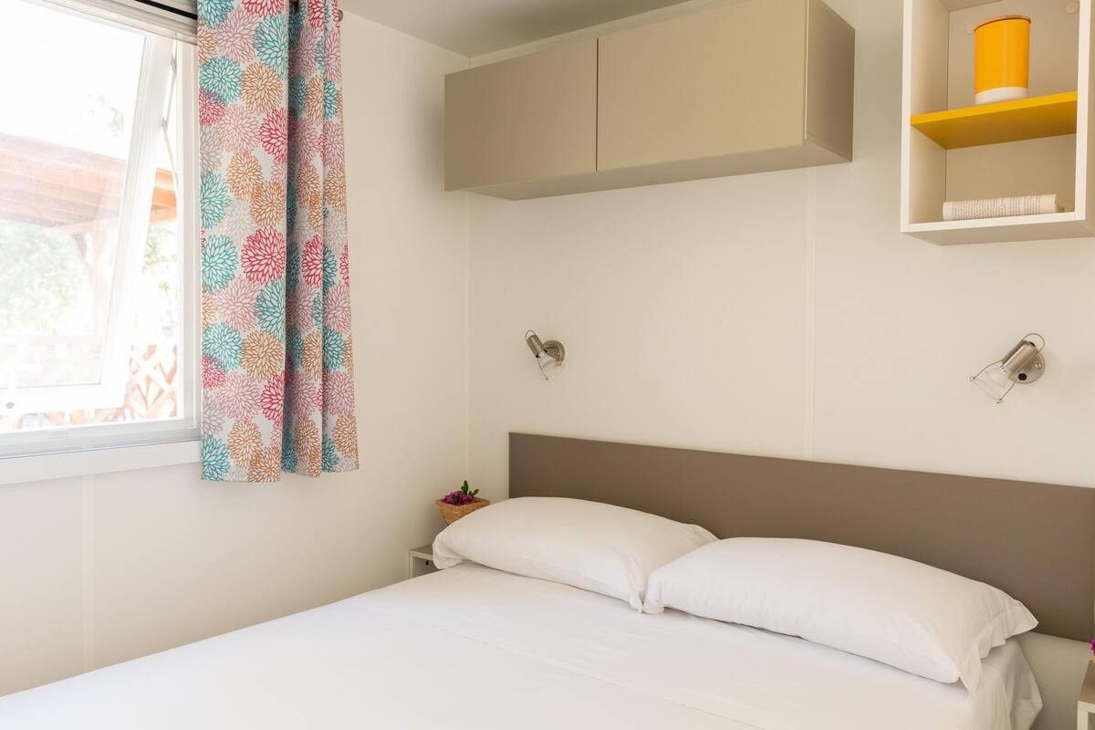 Ein Schlafzimmer mit Bett, einem Fenster mit Vorhängen, einem an der Wand befestigten Regal mit einem gelben Behälter und einem kleinen Tisch mit einem Korb.