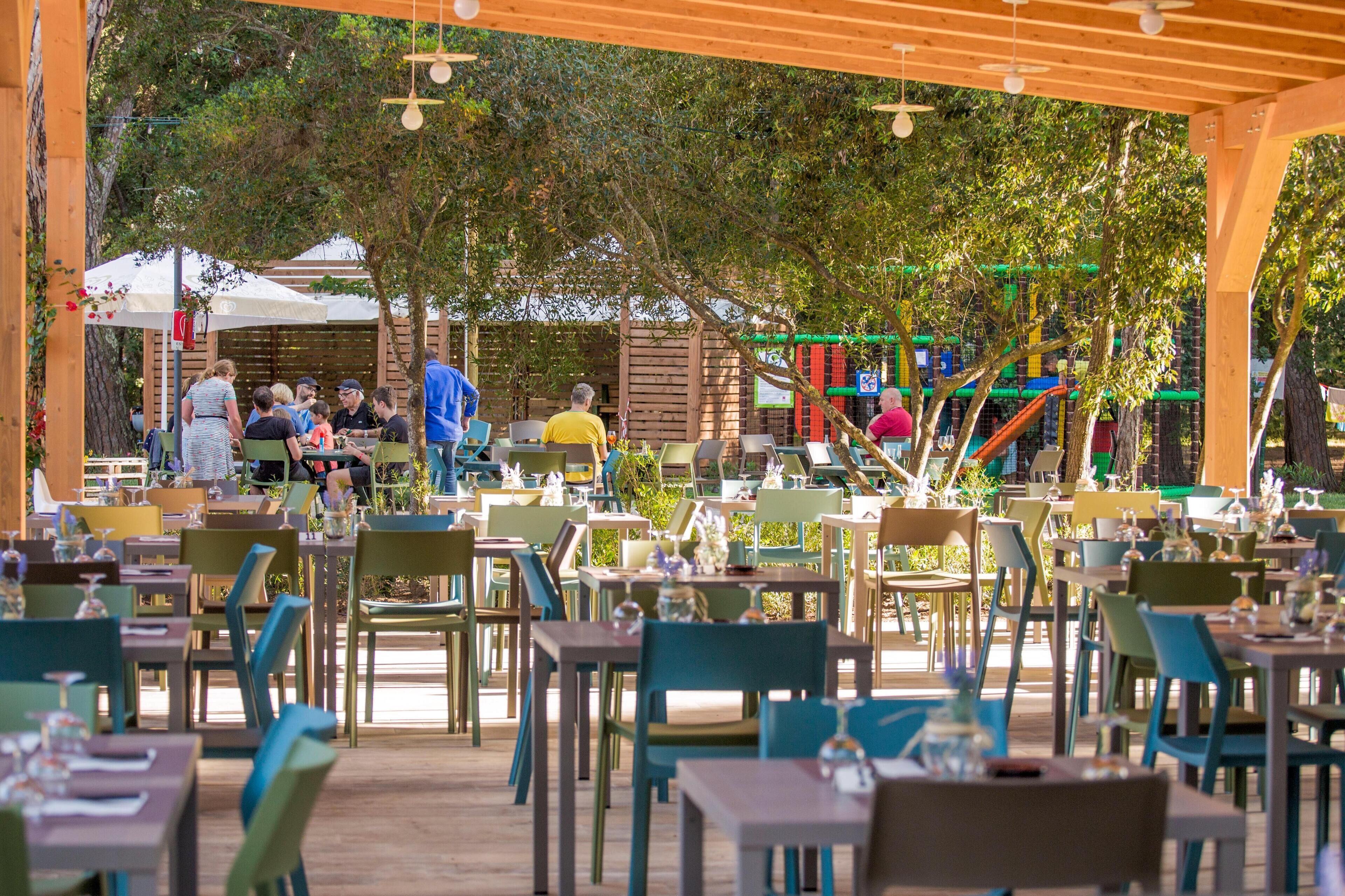 An outdoor dining area with tables and chairs, surrounded by trees and a playground in the background.