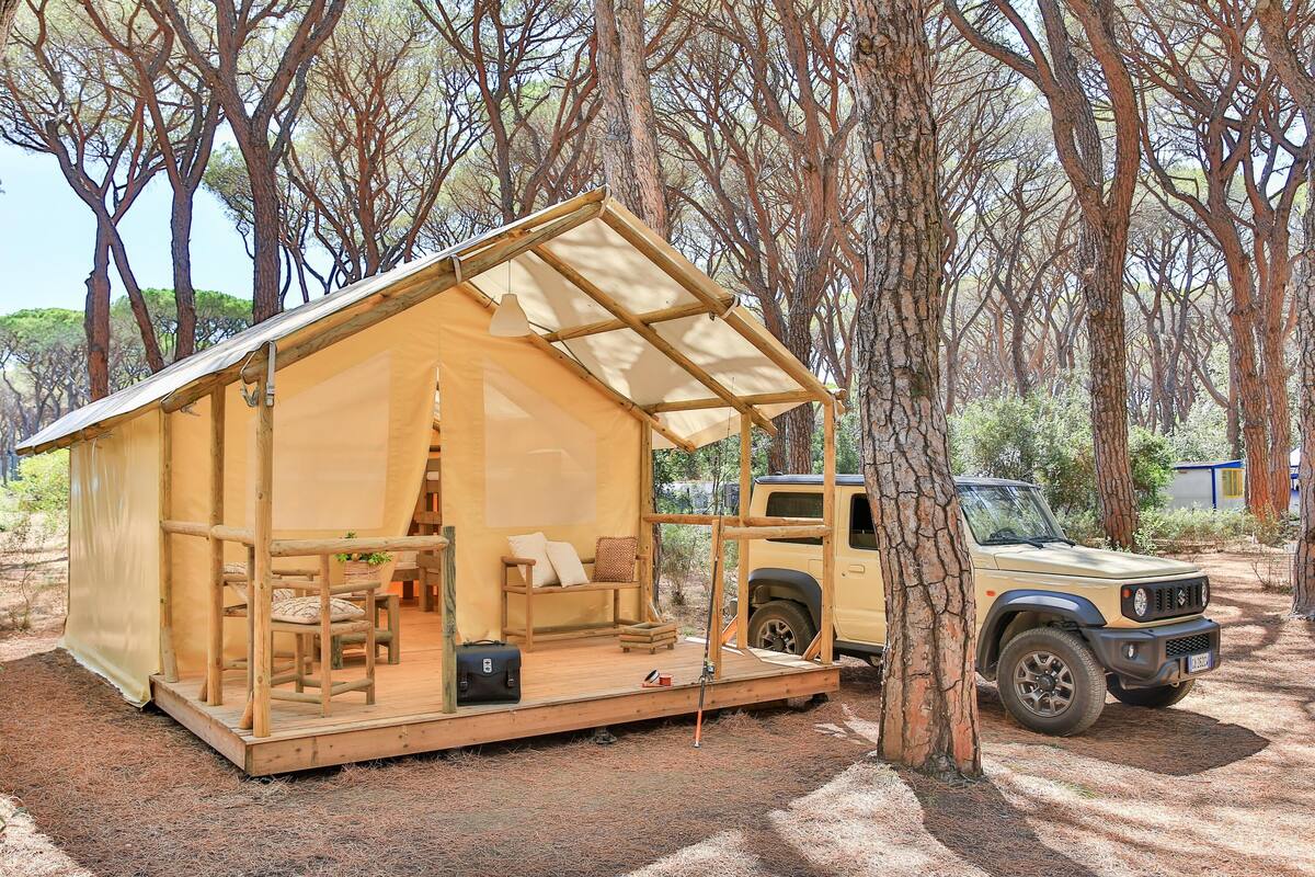 Eine kleine, offene Holzhütte mit überdachter Veranda, einer Couch und einem Esstisch. Ein Fahrzeug ist in der Nähe geparkt.