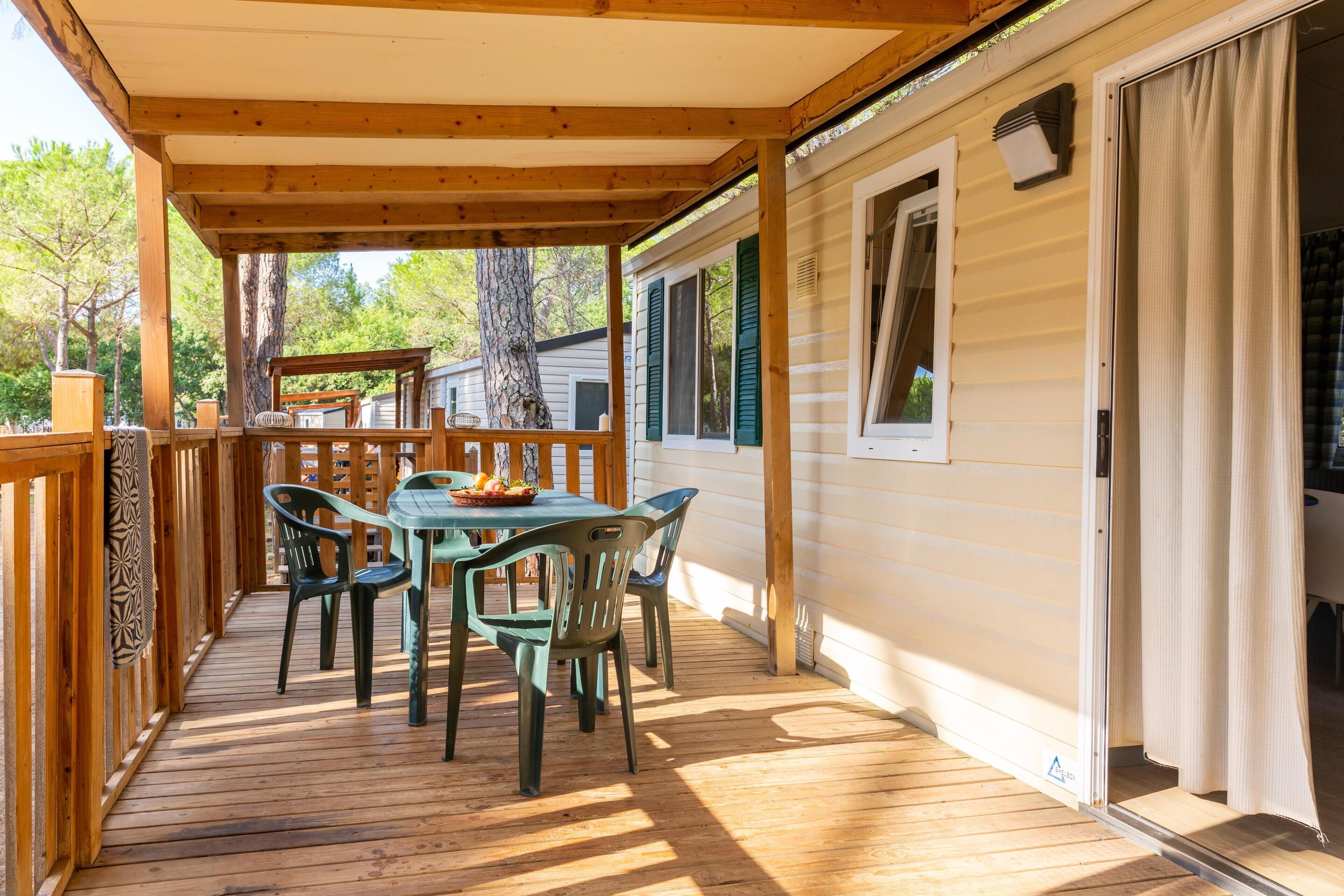 Une terrasse extérieure couverte, dotée d’une table et de chaises, de rampes en bois et offrant une vue sur une maison mobile.