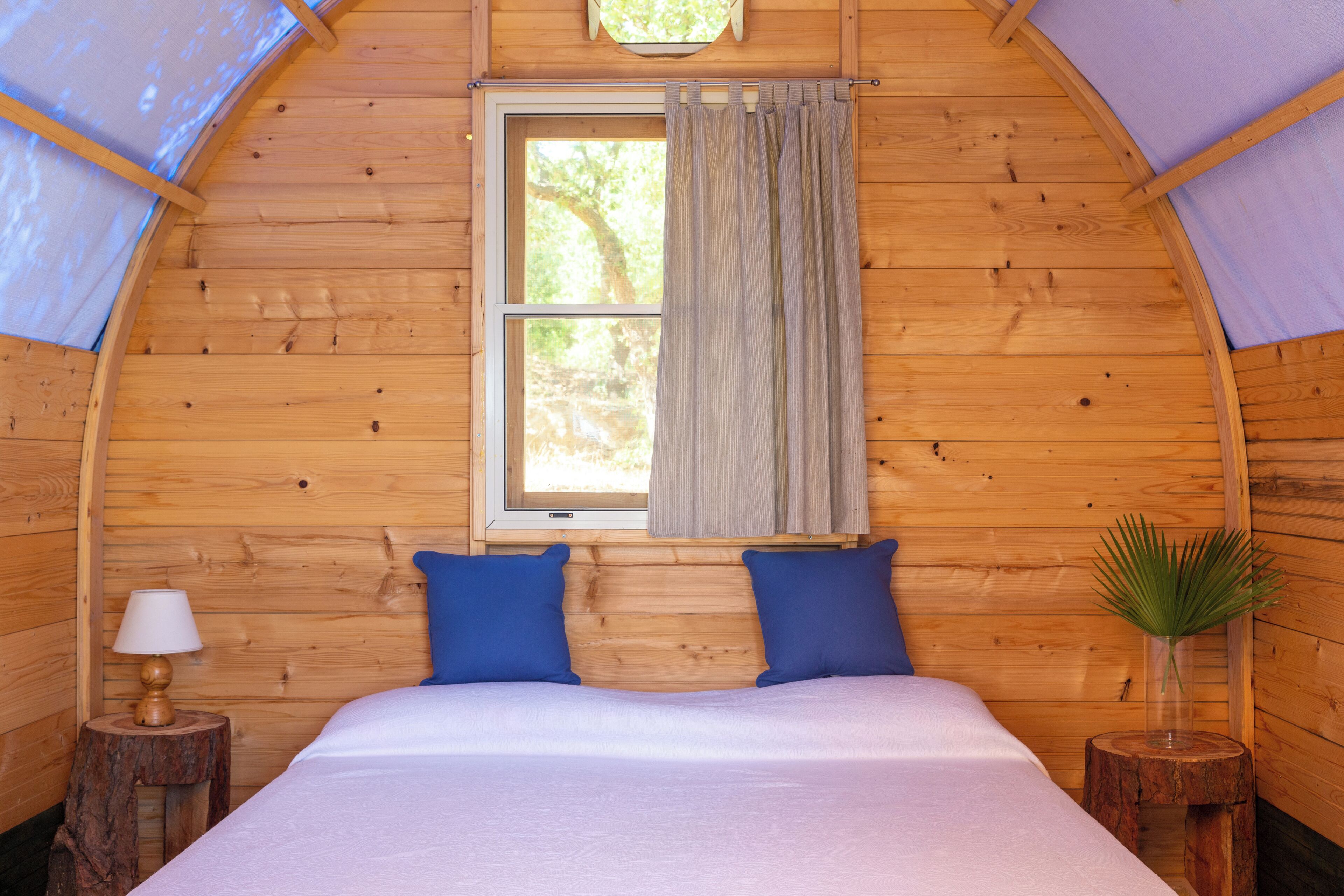 Een houten slaapkamer met een bed, twee kussens, een raam met gordijnen, een lamp en een kleine plant op een houten kruk.