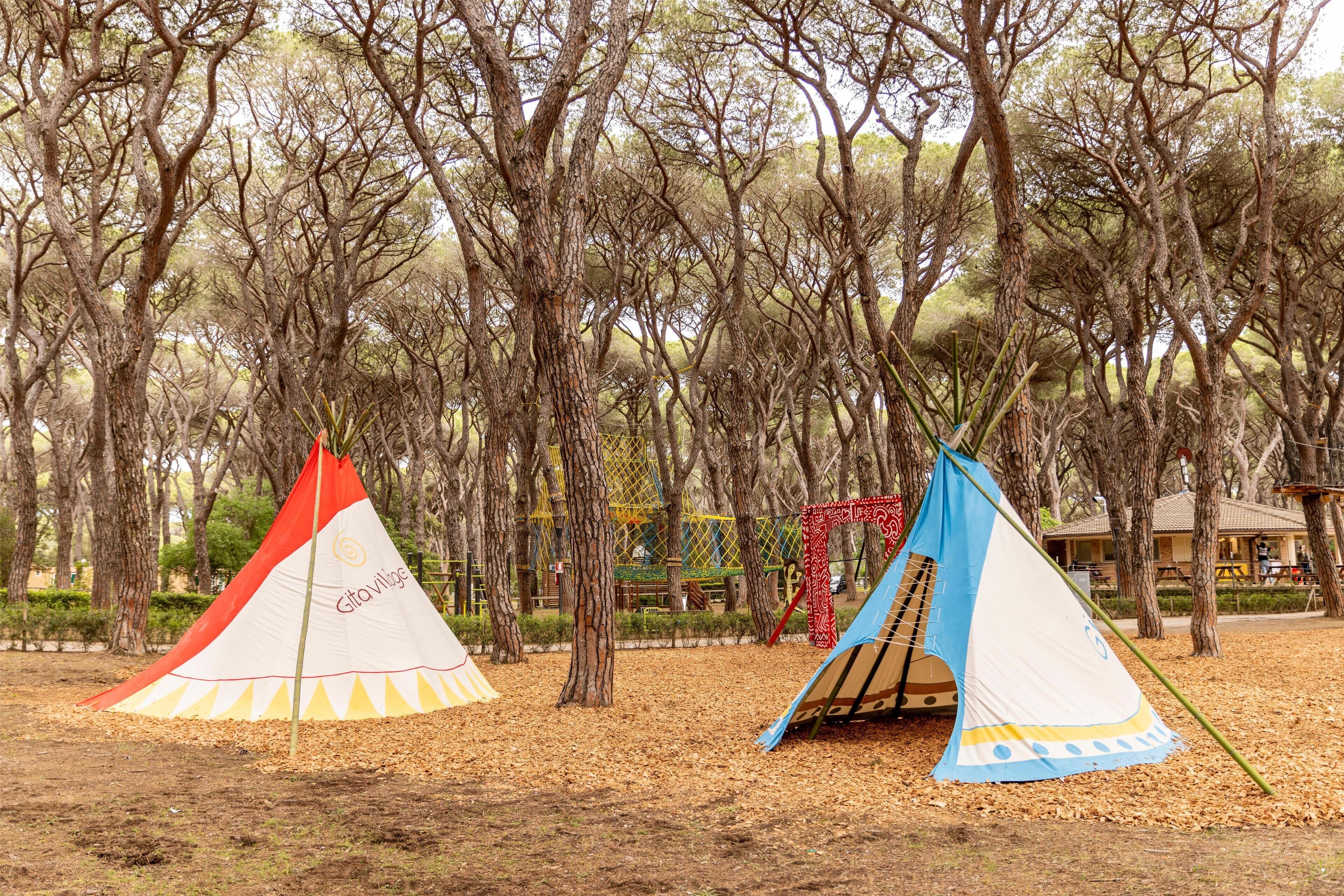 Dos tiendas de campaña grandes en un parque arbolado, con árboles y un parque infantil al fondo.