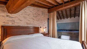 Een slaapkamer met een stenen muur, houten balken in het plafond, een bed, een nachtkastje, een lamp en uitzicht op een keuken door een raam.