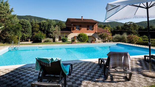 A swimming pool with lounge chairs and an umbrella, surrounded by a house and greenery.