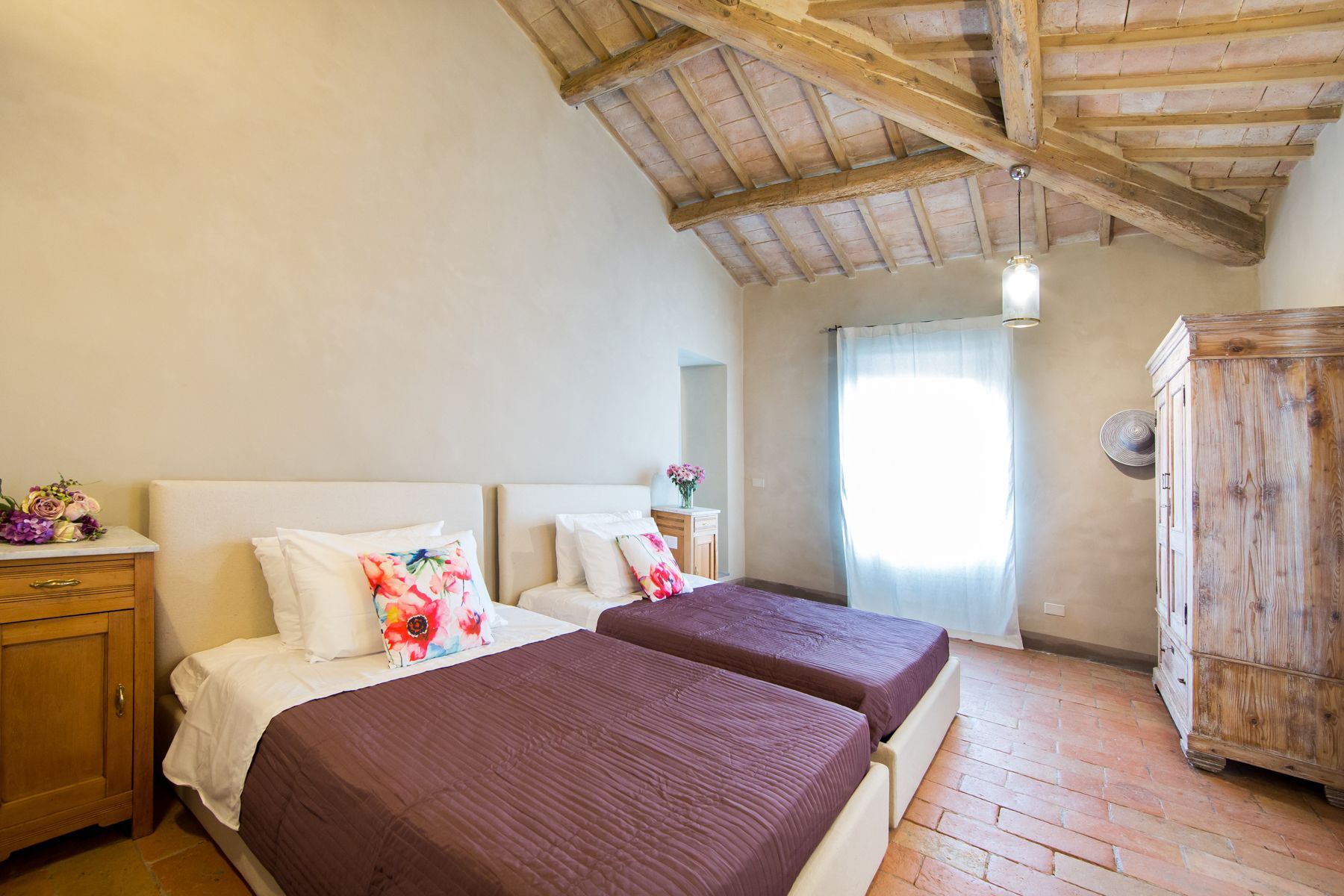 Une chambre avec deux lits, des poutres apparentes en bois au plafond, une armoire en bois et une fenêtre avec des rideaux.
