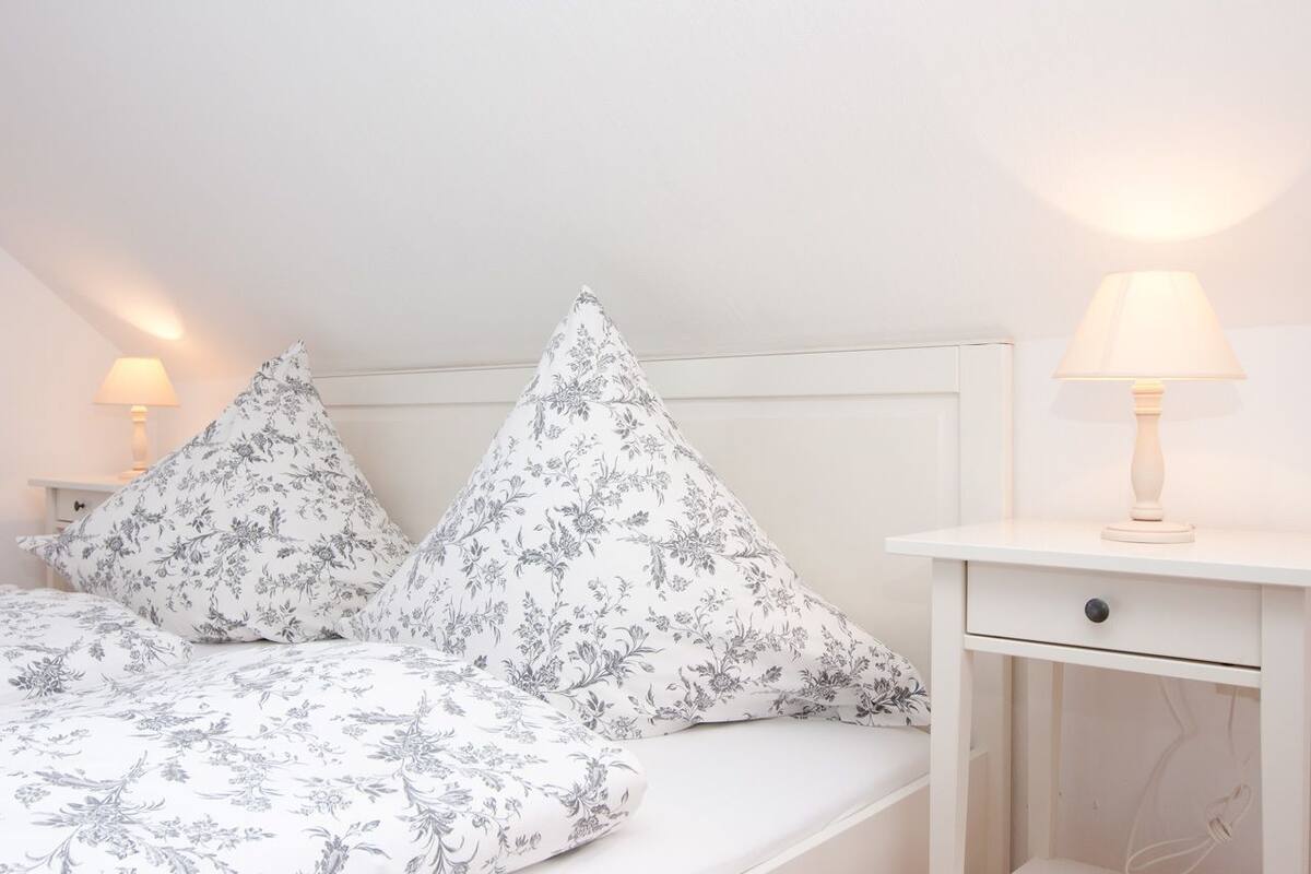 A bedroom with a bed, two pillows, a nightstand, a lamp, and a drawer.