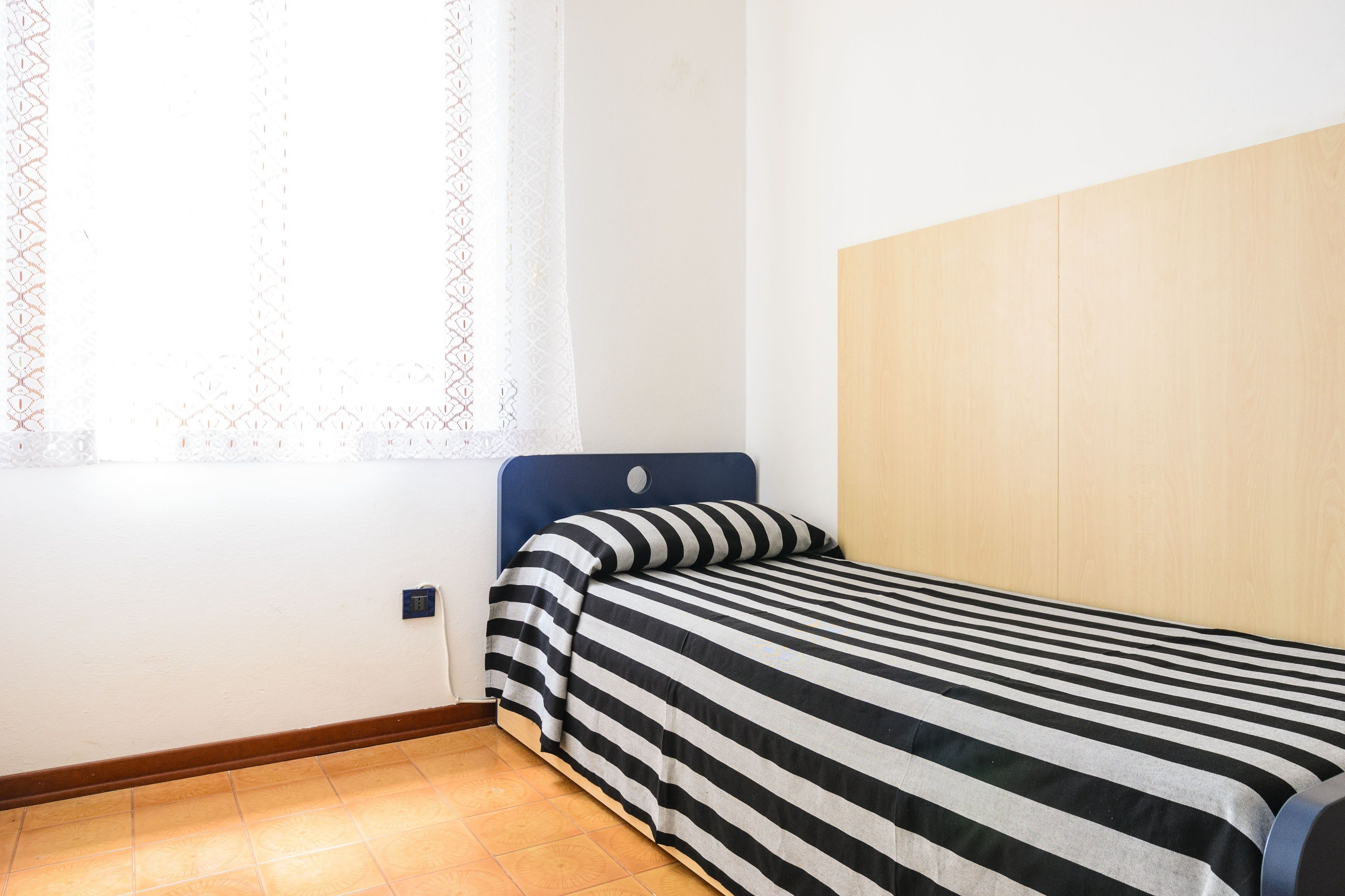 A small, single-bed room with a wooden headboard, a striped bedspread, and a window with a patterned curtain.